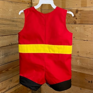 Puede incluir: Un pelele rojo y amarillo con detalles en negro, colgado de una percha blanca. El pelele sin mangas presenta una ancha banda amarilla en la secci&oacute;n media y ribetes negros en los pantalones cortos.