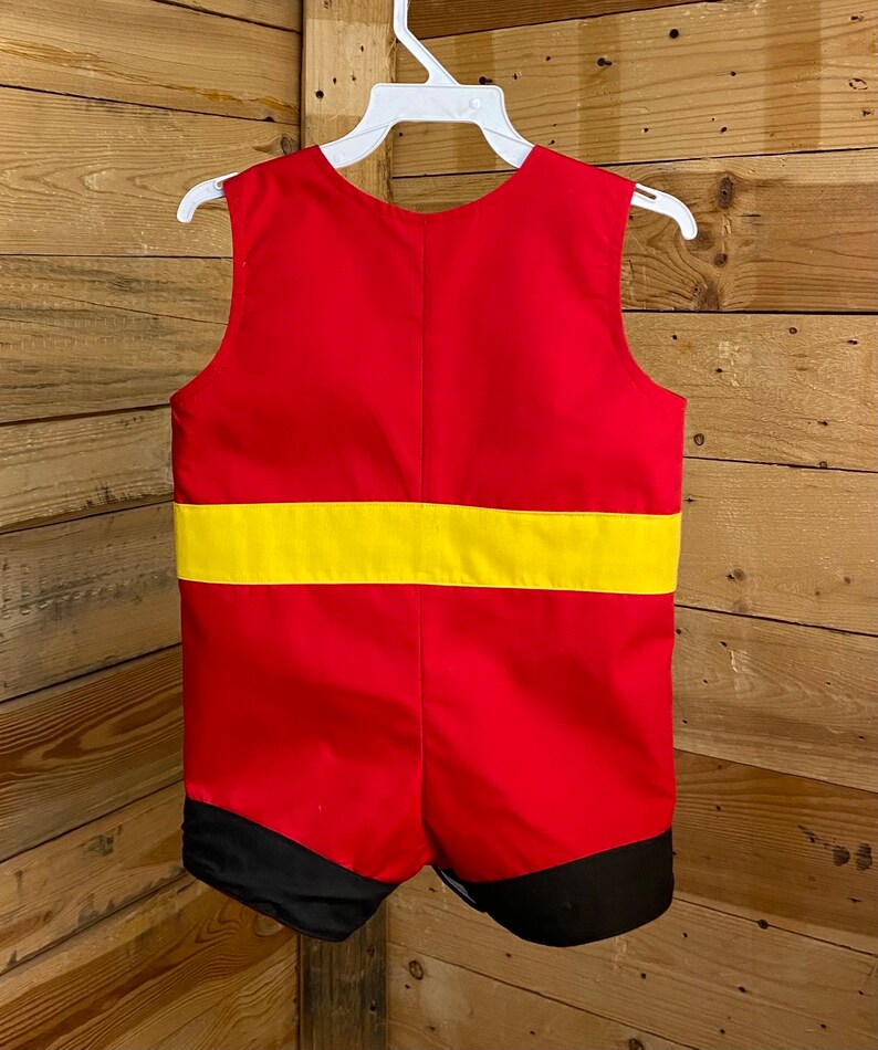 Puede incluir: Un pelele rojo y amarillo con detalles en negro, colgado de una percha blanca. El pelele sin mangas presenta una ancha banda amarilla en la secci&oacute;n media y ribetes negros en los pantalones cortos.