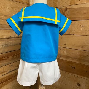 May include: A blue and white sailor-style outfit for a child. The shirt has a wide collar with yellow trim and short sleeves. The shorts are white.