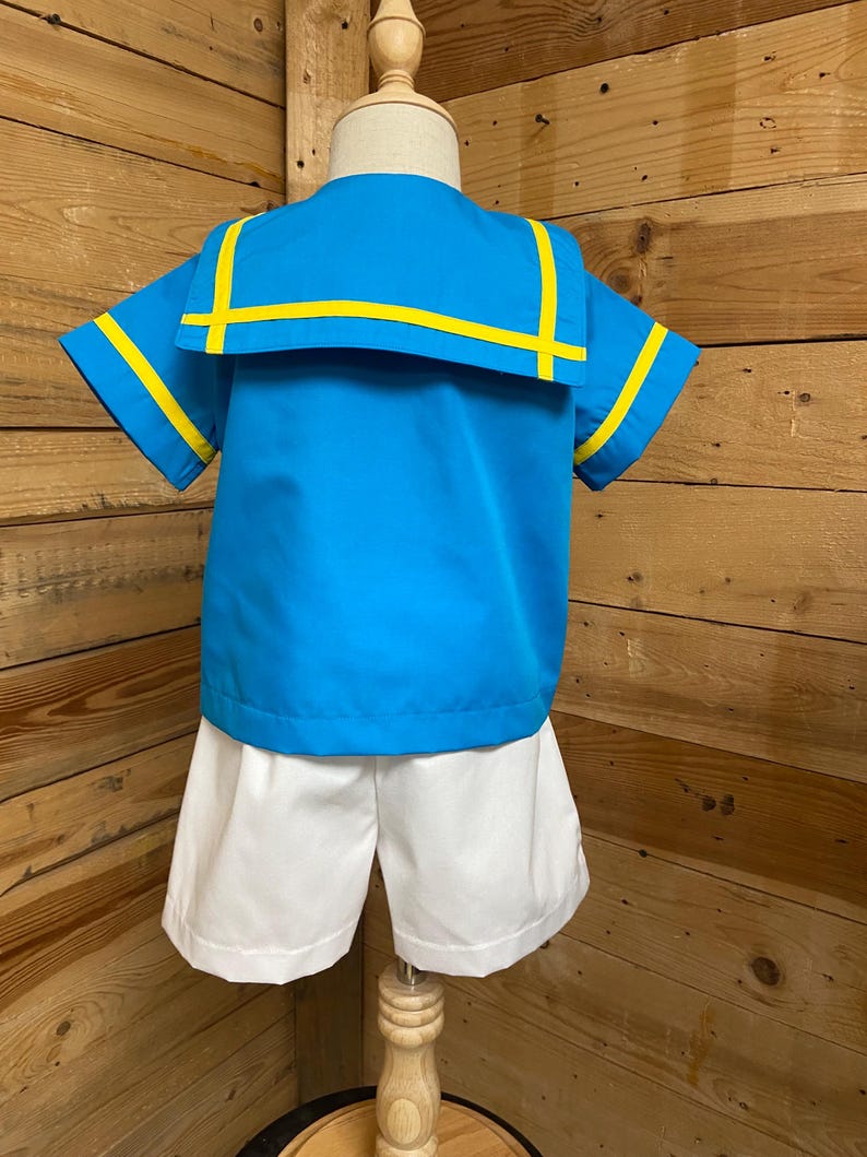 May include: A blue and white sailor-style outfit for a child. The shirt has a wide collar with yellow trim and short sleeves. The shorts are white.