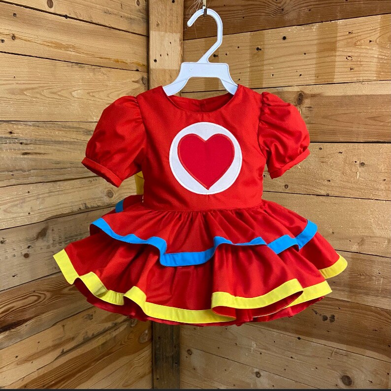 Pode incluir: Um vestido vermelho com um cora&ccedil;&atilde;o branco na frente. O vestido tem uma borda azul e amarela ao redor da parte inferior.