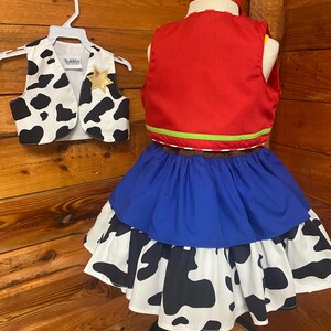 May include: A child's cowboy costume set. The set includes a red vest with green trim, a blue skirt, and a cow-print skirt. A matching cow-print vest with a gold star is on a hanger. The costume is displayed on a mannequin.