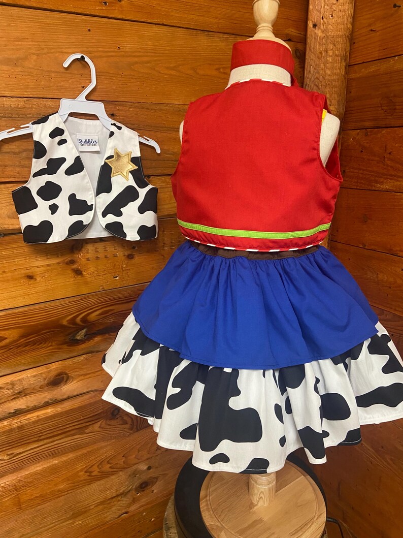 May include: A child's cowboy costume set. The set includes a red vest with green trim, a blue skirt, and a cow-print skirt. A matching cow-print vest with a gold star is on a hanger. The costume is displayed on a mannequin.