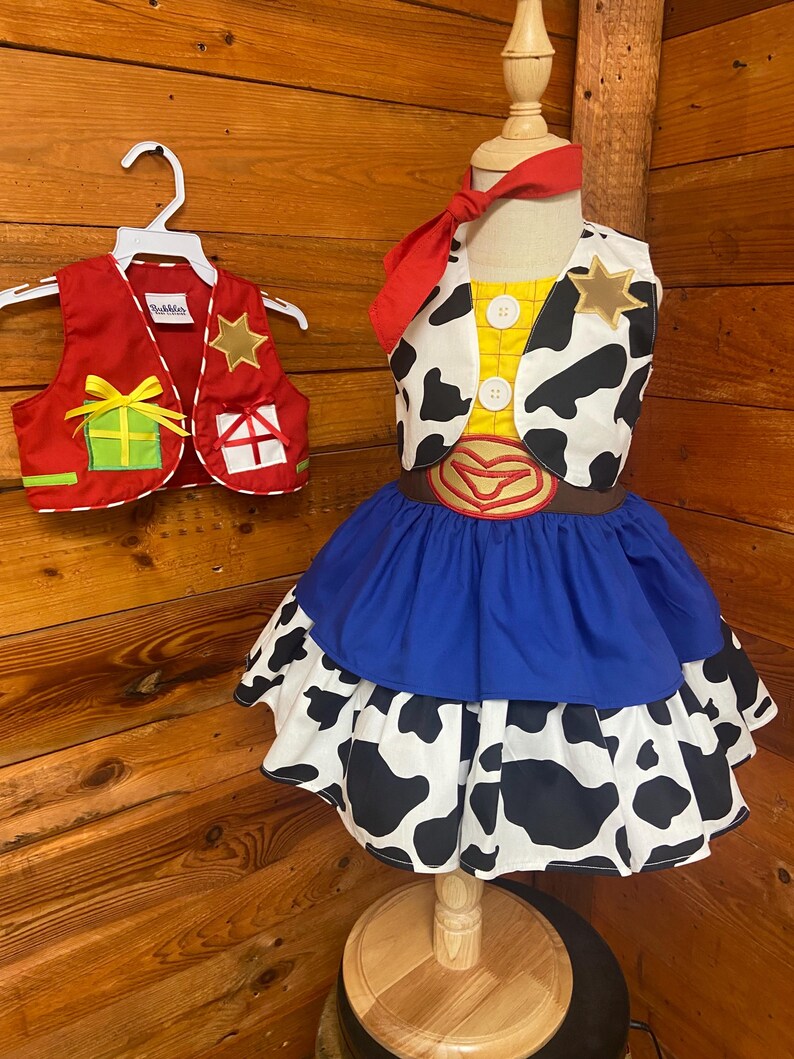 May include: A child's cowgirl costume with a red vest and a dress. The dress has a blue skirt and a cow-print skirt. The vest is red with gold stars and gift box designs. A red bandana is tied around the mannequin's neck.