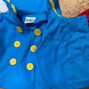 May include: A blue sailor-style jacket with yellow trim and yellow buttons. The jacket has a hood and is made of a soft, lightweight fabric. The label on the jacket reads "Bubbles Baby Clothing."