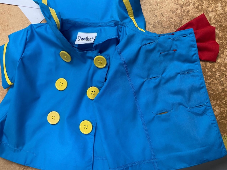 May include: A blue sailor-style jacket with yellow trim and yellow buttons. The jacket has a hood and is made of a soft, lightweight fabric. The label on the jacket reads "Bubbles Baby Clothing."