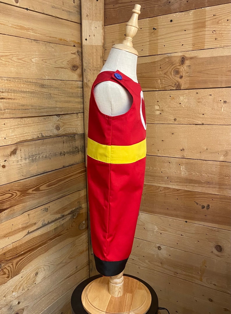 Peut inclure: Un costume rouge avec une bande jaune et un motif de cercle blanc. Le costume a un bouton bleu sur l'&eacute;paule et un ourlet noir. Le costume est pr&eacute;sent&eacute; sur un mannequin.