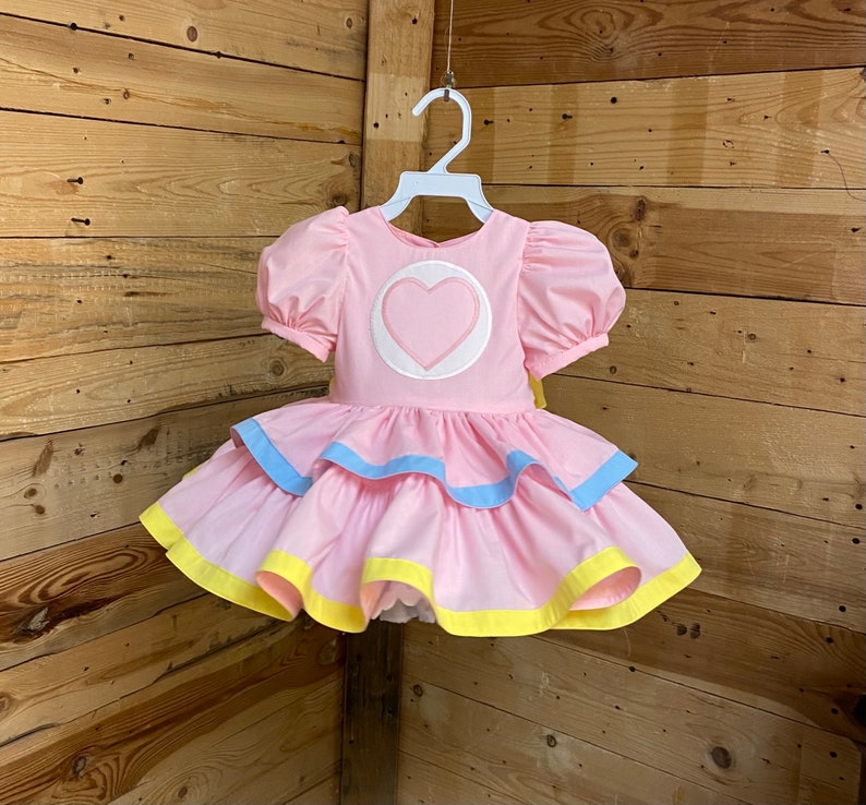 May include: A pink dress with a heart-shaped applique on the bodice and layered skirt. The dress features puff sleeves and is trimmed with blue and yellow accents. It is hanging on a white hanger against a wooden background.