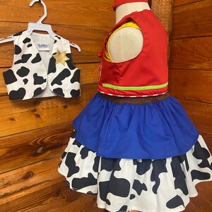 May include: A child's cowgirl costume set. The set includes a red vest with yellow lining, a blue skirt, and a white skirt with black cow spots. A matching vest with a gold star and a red headband are also included.