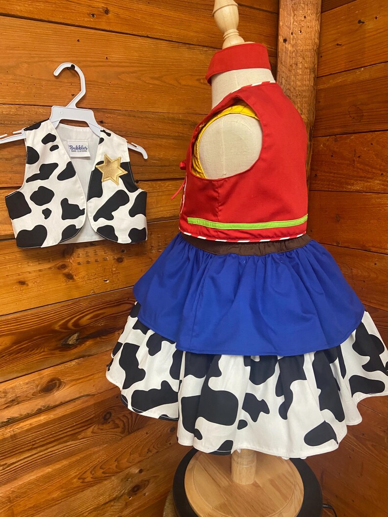 May include: A child's cowgirl costume set. The set includes a red vest with yellow lining, a blue skirt, and a white skirt with black cow spots. A matching vest with a gold star and a red headband are also included.