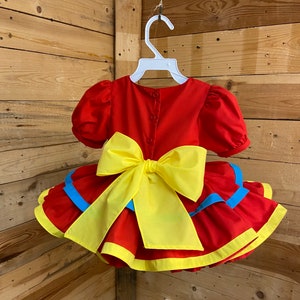 Pode incluir: Vestido vermelho e amarelo com babados e um grande la&ccedil;o amarelo nas costas. O vestido tem detalhes azuis nos babados.