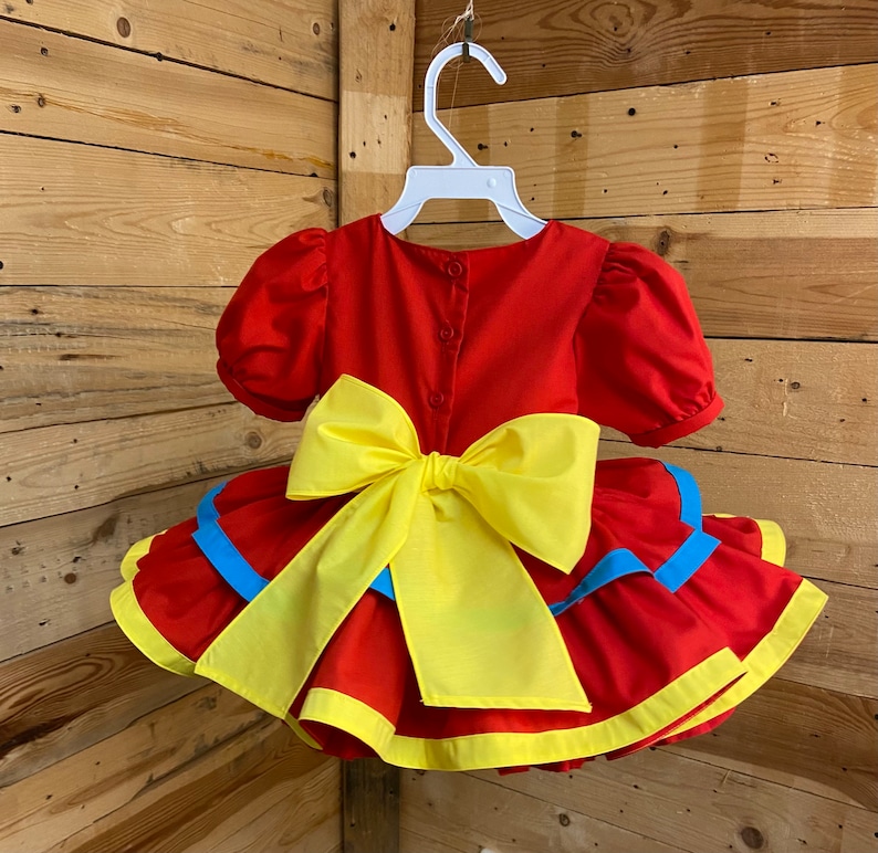 Pode incluir: Vestido vermelho e amarelo com babados e um grande la&ccedil;o amarelo nas costas. O vestido tem detalhes azuis nos babados.