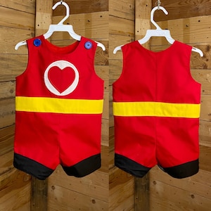 Puede incluir: Romper rojo con una franja amarilla y ribete negro. La parte delantera presenta un c&iacute;rculo blanco con un coraz&oacute;n rojo. Los botones azules aseguran las correas de los hombros. El romper se muestra en una percha blanca sobre un fondo de madera.