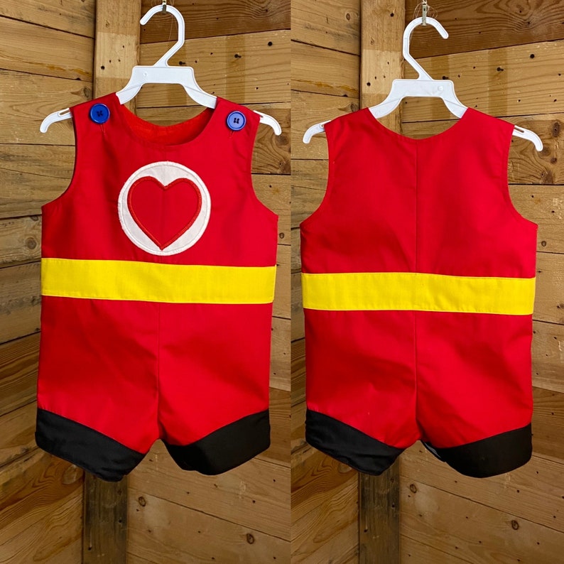 Puede incluir: Romper rojo con una franja amarilla y ribete negro. La parte delantera presenta un c&iacute;rculo blanco con un coraz&oacute;n rojo. Los botones azules aseguran las correas de los hombros. El romper se muestra en una percha blanca sobre un fondo de madera.