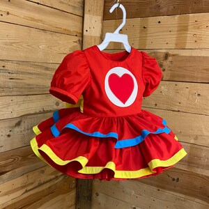 Pode incluir: Um vestido vermelho com um cora&ccedil;&atilde;o branco na frente. O vestido tem uma bainha amarela e azul ao redor da parte inferior.