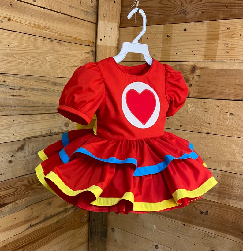 Pode incluir: Um vestido vermelho com um cora&ccedil;&atilde;o branco na frente. O vestido tem uma bainha amarela e azul ao redor da parte inferior.