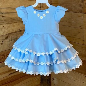 May include: A light blue dress with white lace trim and yellow daisy embroidery. The dress has a ruffled skirt and short sleeves.