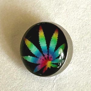 May include: A circular button featuring a vibrant, multi-colored rainbow cannabis leaf graphic set against a solid black background. The design is encased under a smooth, clear, domed surface with a metallic silver-toned rim around the edge.