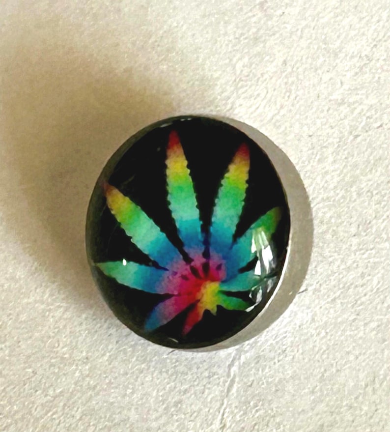 May include: A circular button featuring a vibrant, multi-colored rainbow cannabis leaf graphic set against a solid black background. The design is encased under a smooth, clear, domed surface with a metallic silver-toned rim around the edge.