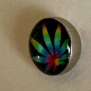 May include: A circular button featuring a glossy, domed surface with a vibrant, multi-colored tie-dye style cannabis leaf graphic set against a solid black background, encased in a smooth, polished silver-toned metal rim.
