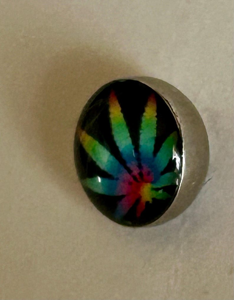 May include: A circular button featuring a glossy, domed surface with a vibrant, multi-colored tie-dye style cannabis leaf graphic set against a solid black background, encased in a smooth, polished silver-toned metal rim.