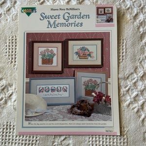 May include: A book cover titled "Sweet Garden Memories" featuring framed floral cross-stitch designs. The cover includes the text "Shawn Mory McMillion's" and a quote. A white hat and a basket are in the foreground.