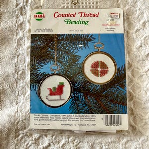 May include: A craft kit for counted thread beading, featuring two designs. The kit includes glass beads, cotton fabric, embroidery floss, a needle, and a gold metallic thread. The designs are displayed in 2.5-inch round frames. The package has the text "Counted Thread Beading".