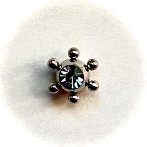 May include: A silver-colored, star-shaped stud earring with a central, faceted, dark-colored gem. The earring has six small, round, silver-colored balls evenly spaced around the central gem. The background is a soft, white, textured surface.