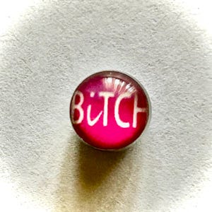 May include: A small, round, pink charm with the word "B*TCH" written in white letters.