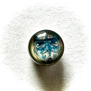 May include: A small, round, silver-colored button with a glass-like top. The button features a blue and white image of a Stormtrooper helmet from Star Wars. The image is centered on the button.