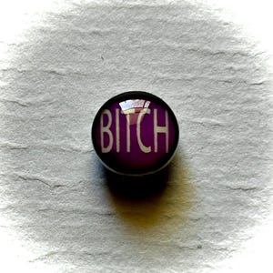 May include: A round, purple button with the word "BITCH" in white, bold letters. The button has a shiny, reflective surface and is set against a textured, gray background.