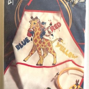 May include: A Vogart Crafts play smock kit for embroidery or ballpoint painting in original packaging. The white smock features a printed giraffe with a monkey rider, paint buckets, and the words BLUE, RED, and YELLOW. Includes embroidery hoop.