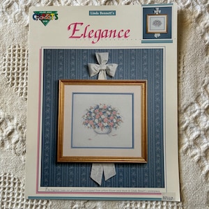May include: A cross-stitch pattern titled "Elegance" by Linda Bennett, featuring a floral arrangement in a gold frame. The design is set against a blue patterned background with a white bow. The text "Colors" and "97117" are also visible.