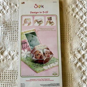 May include: Sizzix Bigz die for 3D design. The package features a sample of a 3D photo display with the text "Many Faces of Aubrey". The box also includes the text "Design in 3-D!" and "Extra Long".