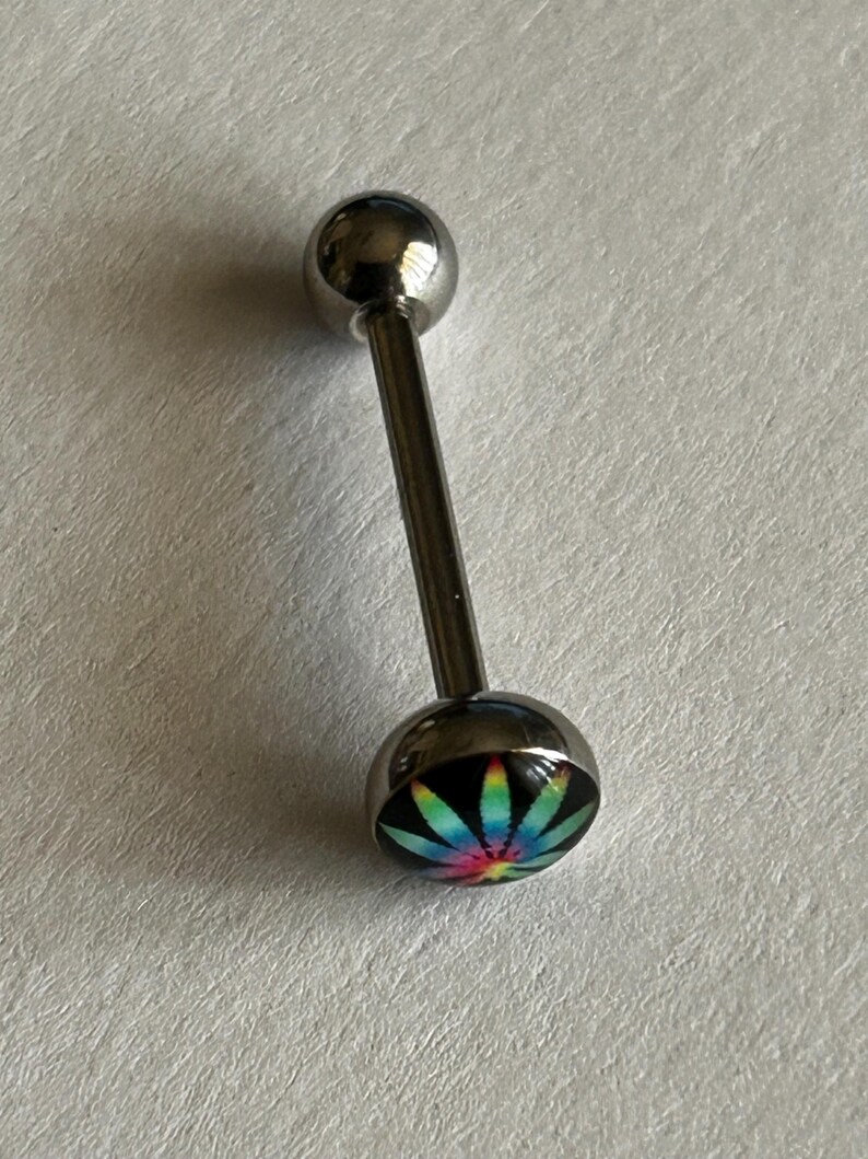 May include: A silver-toned barbell body jewelry piece featuring a straight post with a smooth spherical top and a bottom dome-shaped end. The bottom dome displays a vibrant, multi-colored tie-dye leaf graphic set against a solid black background.