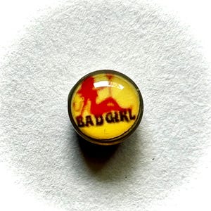 May include: A round, yellow button with a red silhouette of a person and the words "BAD GIRL" in black. The button has a shiny, glass-like surface and a gold-colored rim.