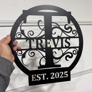 Op de afbeelding: Zwart metalen bord met een decoratief krulontwerp en de letter "T" in het midden. De naam "TRAVIS" staat onder de letter en "EST. 2025" staat onder de naam.