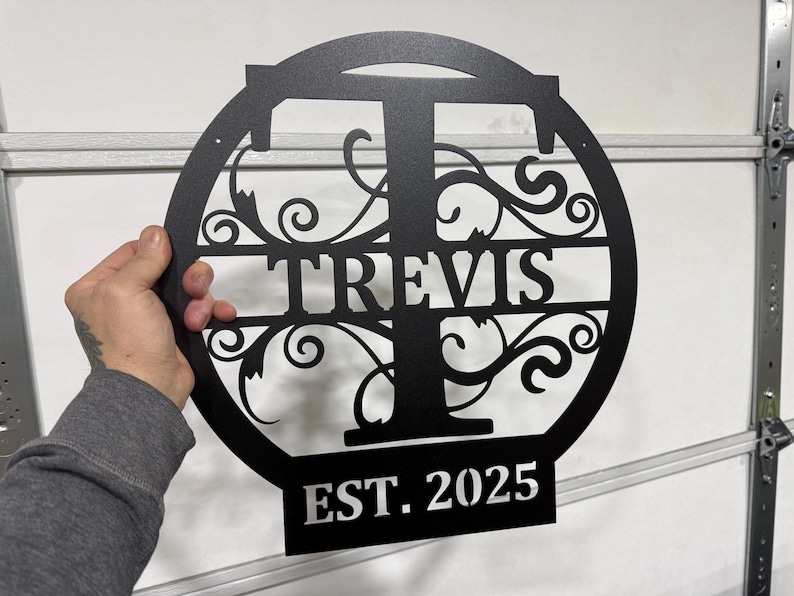 Op de afbeelding: Zwart metalen bord met een decoratief krulontwerp en de letter "T" in het midden. De naam "TRAVIS" staat onder de letter en "EST. 2025" staat onder de naam.