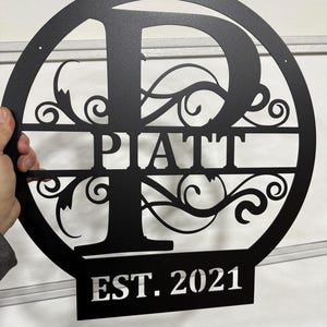 Op de afbeelding: Een zwart metalen rond bord met een grote letter "P" in het midden, omringd door decoratieve krulletjes. De naam "PATT" staat onder de letter en "EST. 2021" staat onder de naam.