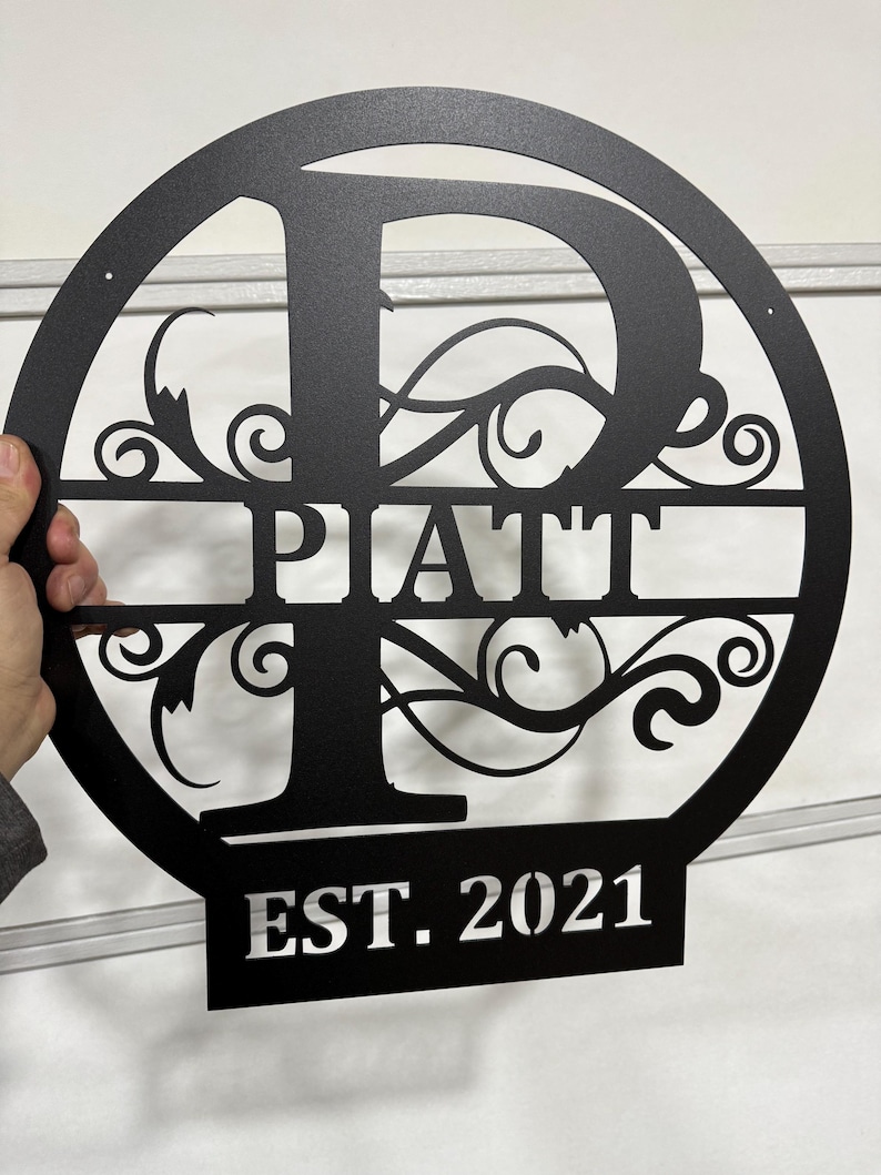 Op de afbeelding: Een zwart metalen rond bord met een grote letter "P" in het midden, omringd door decoratieve krulletjes. De naam "PATT" staat onder de letter en "EST. 2021" staat onder de naam.