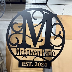 Op de afbeelding: Een rond zwart metalen bord met een grote letter M in het midden, omringd door decoratieve krulletjes. Het bord bevat ook de tekst "McEowen Patio EST. 2024".