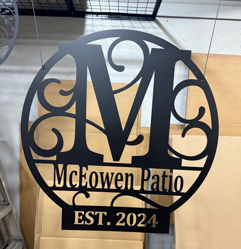 Op de afbeelding: Een rond zwart metalen bord met een grote letter M in het midden, omringd door decoratieve krulletjes. Het bord bevat ook de tekst "McEowen Patio EST. 2024".