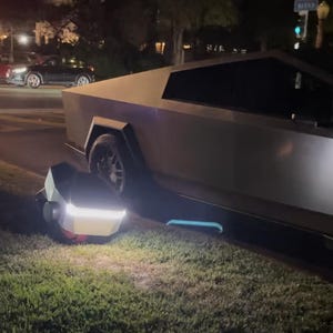 Puede incluir: Un vehículo plateado de aspecto futurista estacionado sobre hierba por la noche. Un objeto blanco y rojo con una luz brillante está posicionado cerca de la parte delantera del vehículo. Otros coches son visibles en el fondo.