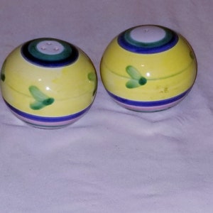 May include: A pair of yellow ceramic salt and pepper shakers with green and blue painted designs. The shakers are round and have a white top.