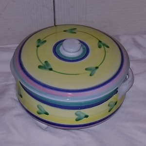 May include: A yellow ceramic casserole dish with a lid. The dish has a blue, pink, and green striped design with green leaf accents. The lid has a white knob.