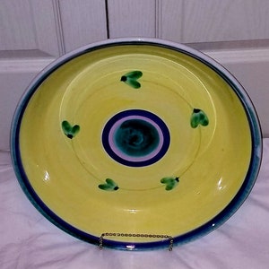May include: A yellow ceramic plate with a blue and green design. The plate has a concentric circle pattern with a green centre and blue and pink rings. There are four green leaf designs around the centre circle.