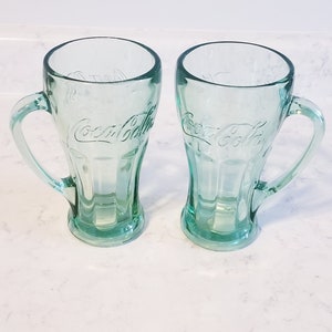 Puede incluir: Dos tazas de Coca-Cola de vidrio verde con asas. Las tazas son transparentes y tienen el logotipo de Coca-Cola en relieve en la parte delantera.