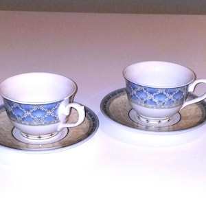 May include: Two white teacups with blue and gold patterned designs on the side. Each cup is on a matching saucer with a similar pattern.