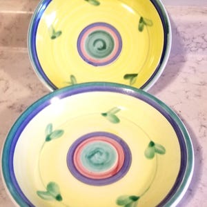 May include: Two yellow ceramic plates with blue, green, and pink painted designs. The plates have a concentric circle design with a swirl in the center and green leaf accents.
