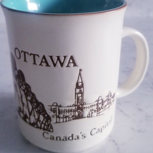 May include: White ceramic mug with a turquoise blue rim. The mug features a brown illustration of the Parliament Buildings in Ottawa, Canada, with the text "OTTAWA" and "Canada's Capital" printed below the image.